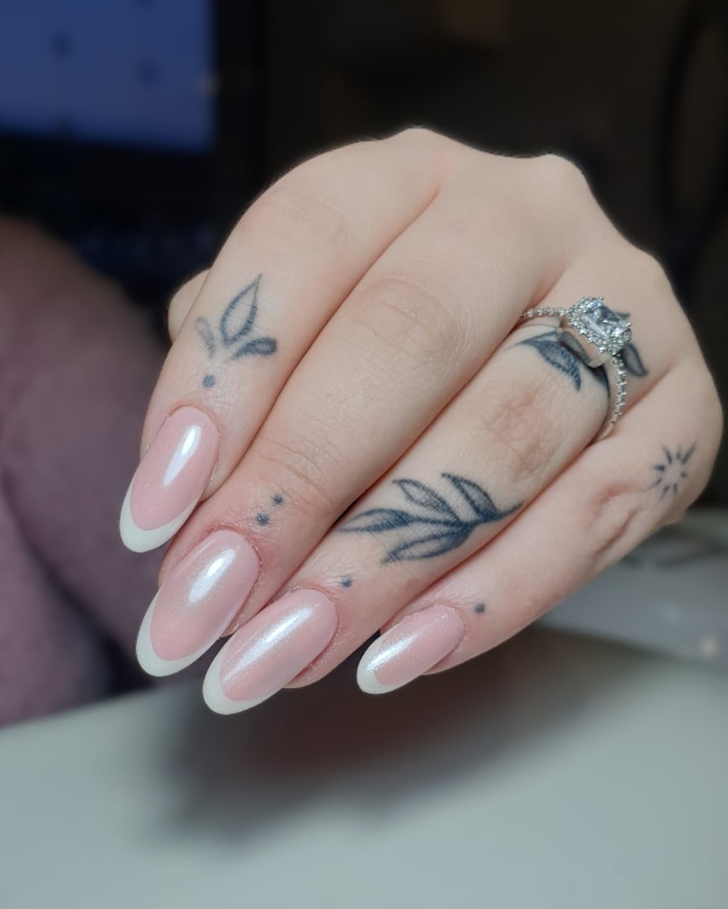 Dłoń z owalnymi paznokciami w stylu french z perłowym wykończeniem, ozdobiona tatuażami i pierścionkiem z dużym kamieniem.