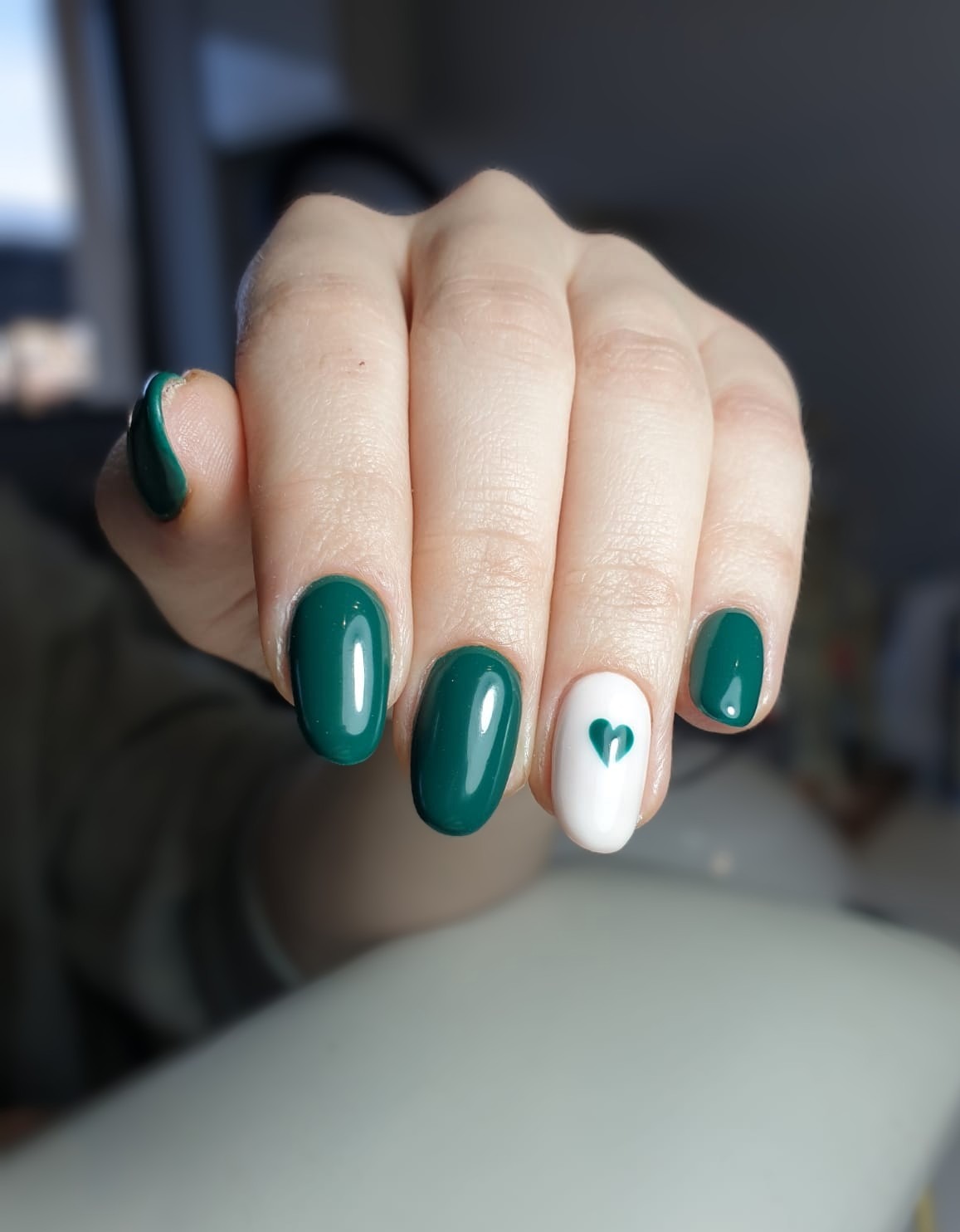 Dłoń z pomalowanymi paznokciami: cztery w odcieniu butelkowej zieleni, jeden biały z małym zielonym serduszkiem. Krótkie, owalne paznokcie.