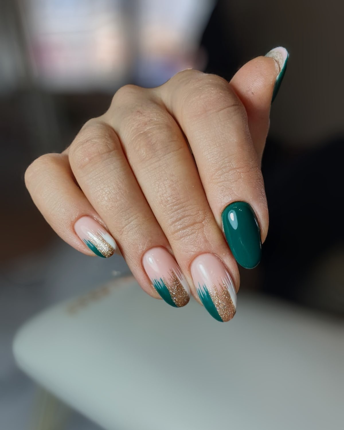 Dłoń z zielono-złotym zdobieniem paznokci w stylu french, na jasnoróżowym tle, jeden paznokieć pomalowany na jednolity ciemnozielony kolor.