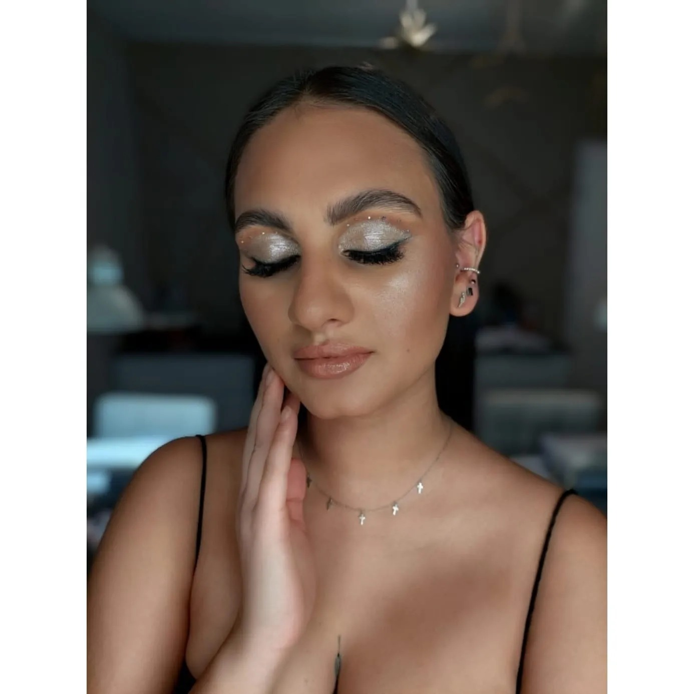 Portret kobiety z zamkniętymi oczami, srebrzystym makijażem z brokatem i drobnymi kryształkami na powiekach, podkreślonymi rzęsami oraz błyszczącymi ustami, dłoń delikatnie dotyka policzka.