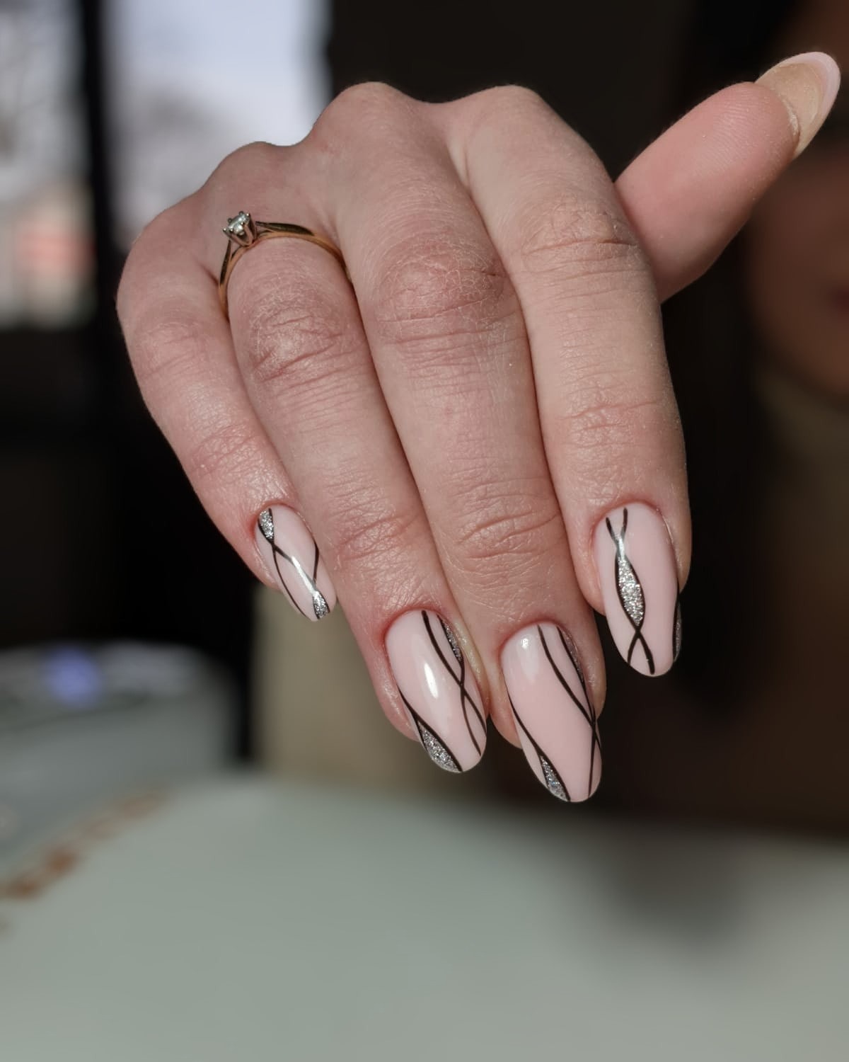 Delikatny manicure hybrydowy w odcieniach pudrowego różu z czarnymi, asymetrycznymi liniami i srebrnymi akcentami brokatu, prezentowany na dłoni z pierścionkiem zaręczynowym.