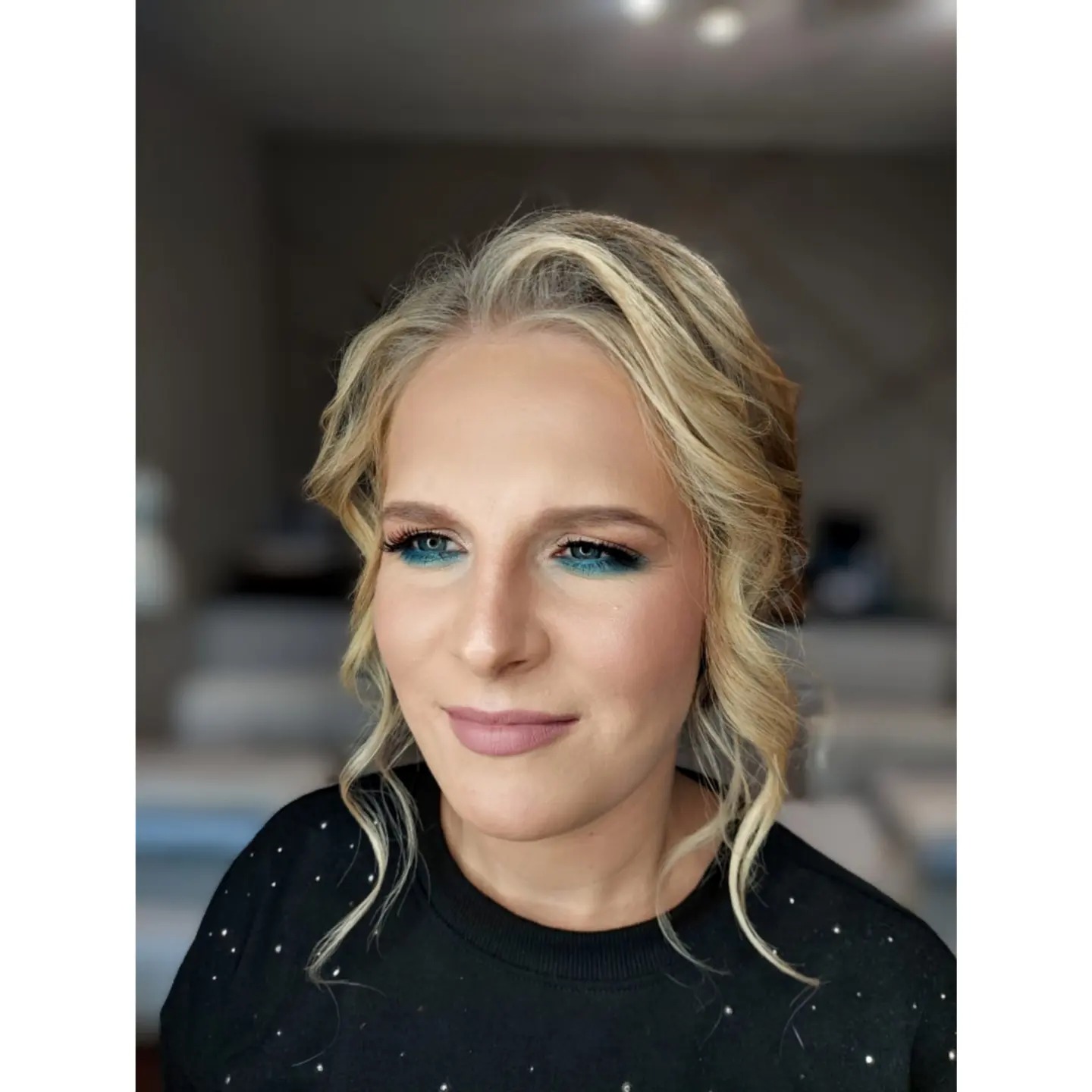 Portret kobiety z profesjonalnym makijażem oczu w odcieniach turkusu i różu, delikatne fale blond włosów, matowa pomadka w odcieniu nude, czarna bluzka z drobnymi zdobieniami.
