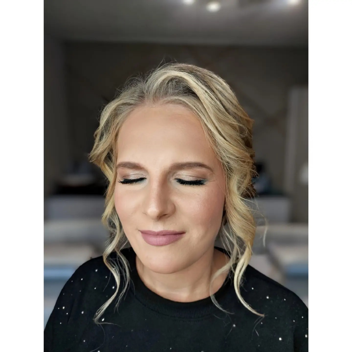 Portret kobiety z zamkniętymi oczami, prezentujący delikatny makijaż dzienny z subtelnym cieniem do powiek, wydłużonymi rzęsami i matową pomadką w odcieniu nude, z blond lokami opadającymi na ramiona.