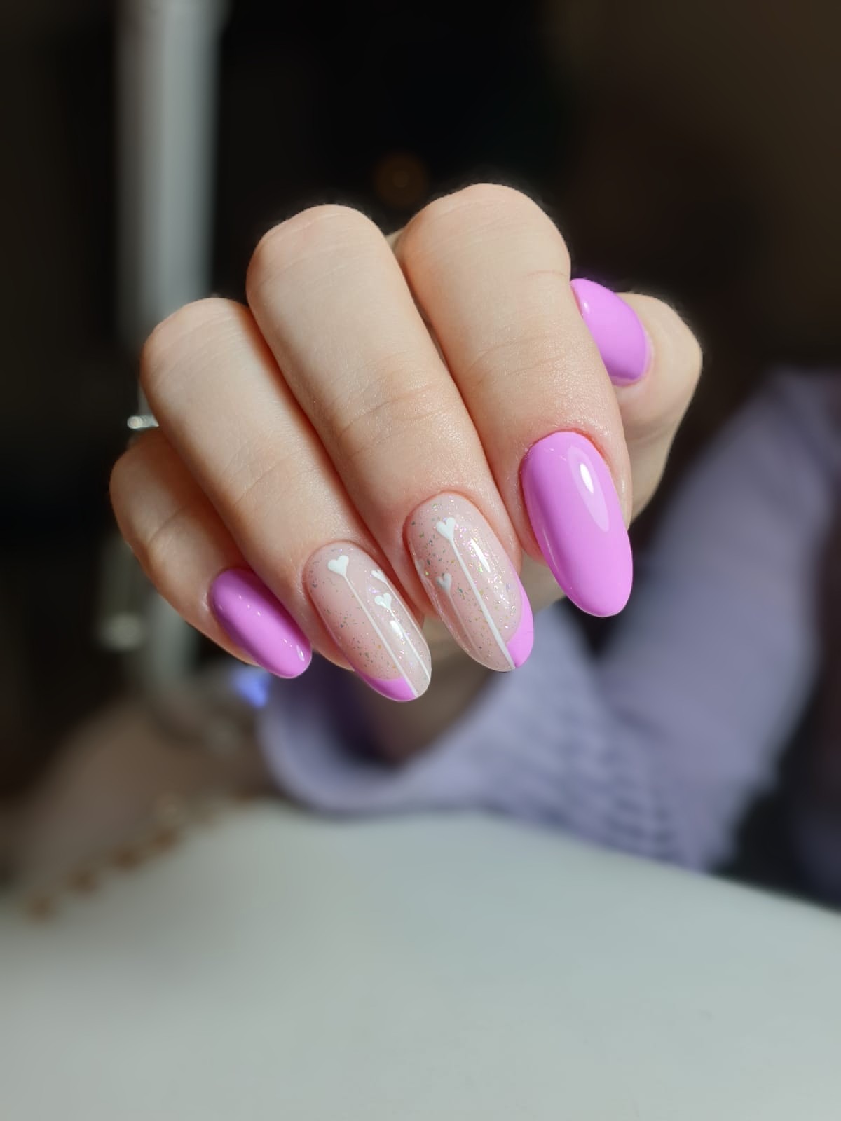 Dłoń z manicure hybrydowym: dwa paznokcie w kolorze liliowym, pozostałe z jasnoróżową bazą, białymi serduszkami i liliową końcówką, wykończone błyszczącym pyłkiem.