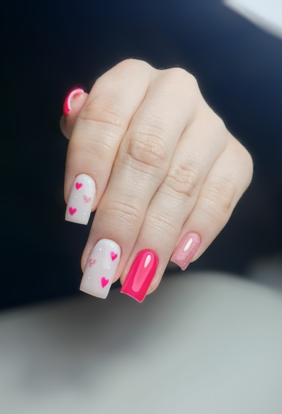 Dłoń z kwadratowymi paznokciami: dwa białe z różowymi serduszkami i kropkami, jeden różowy i jeden brokatowy, na rozmytym ciemnym tle.