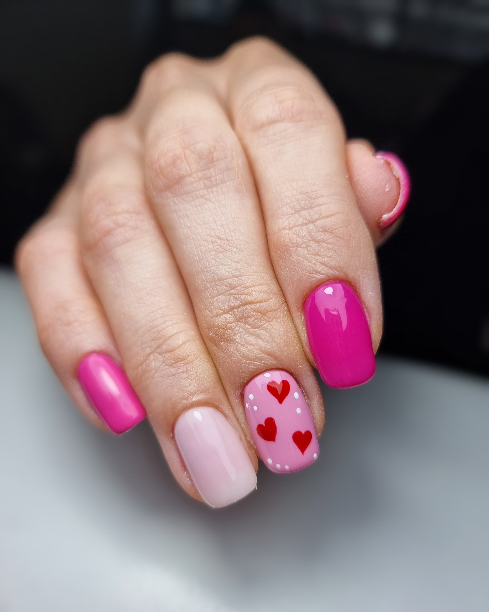Dłoń z pomalowanymi paznokciami w odcieniach różu, jeden paznokieć ozdobiony czerwonymi sercami i białymi kropkami.