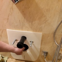 Dirty Water - Instalacja stelaża podtynkowego WC na marmurowej ścianie, widoczny fragment muszli, rurka odpływowa i dłoń instalatora przy otworze.