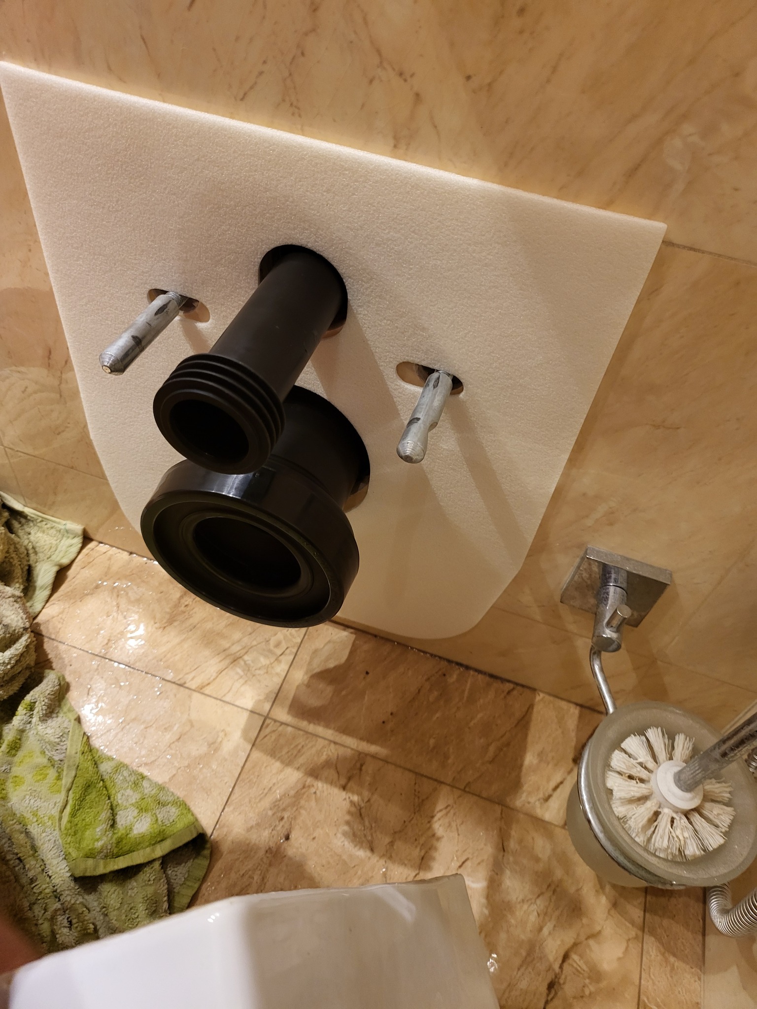 Instalacja odpływu toalety podtynkowej z widocznymi elementami mocującymi, rurami kanalizacyjnymi i fragmentem toalety w tle. Widoczna również szczotka do WC.