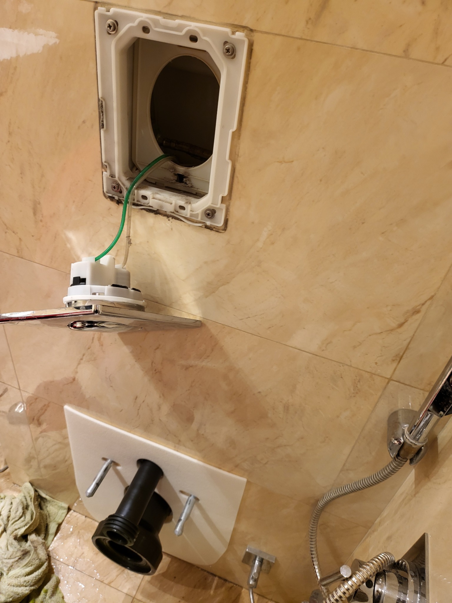 Instalacja podtynkowa WC: biała rama montażowa z widocznymi elementami mocującymi, zielony przewód, chromowany przycisk spłukujący na tle beżowych płytek ceramicznych w łazience.