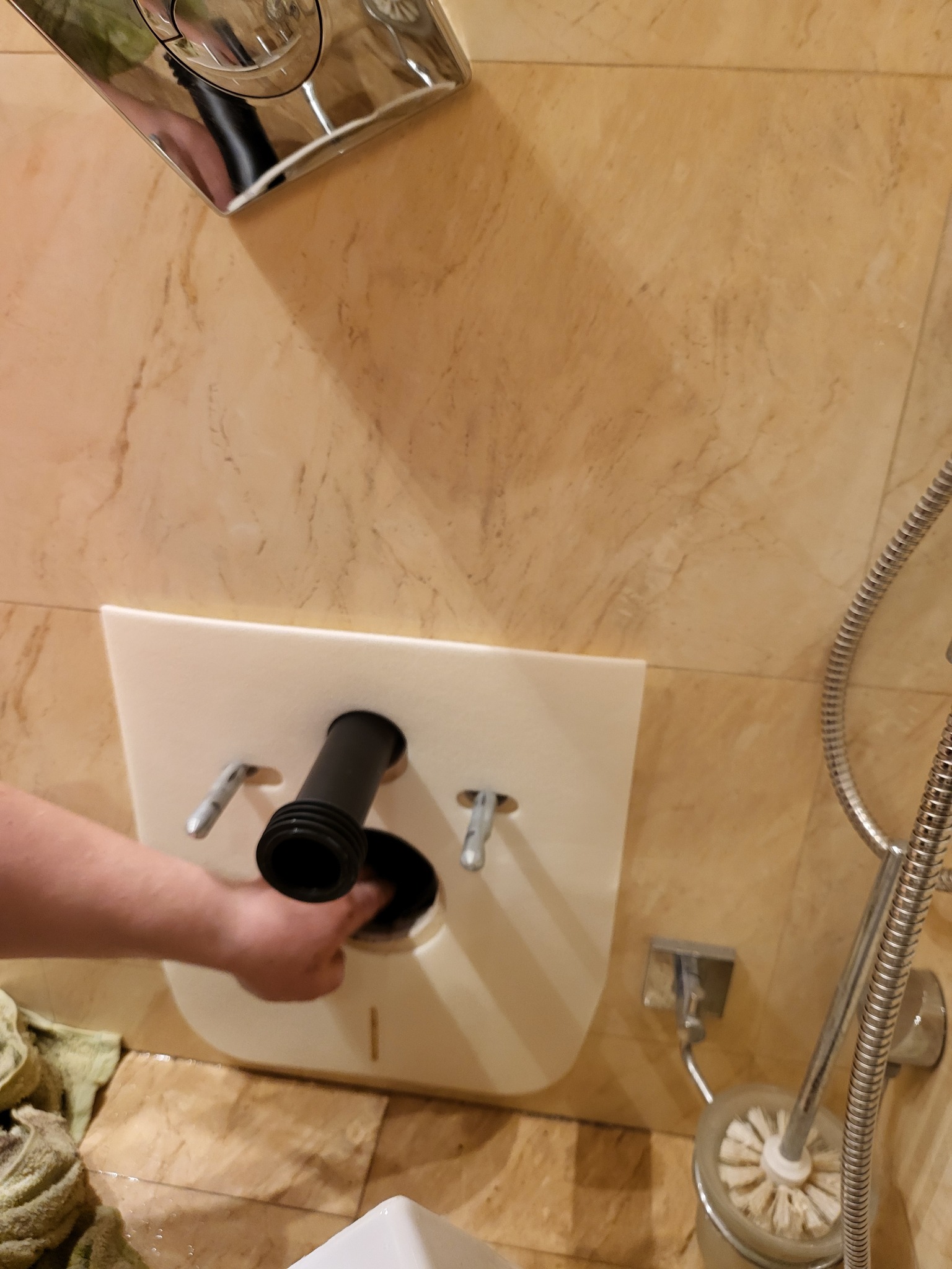 Instalacja stelaża podtynkowego WC na marmurowej ścianie, widoczny fragment muszli, rurka odpływowa i dłoń instalatora przy otworze.