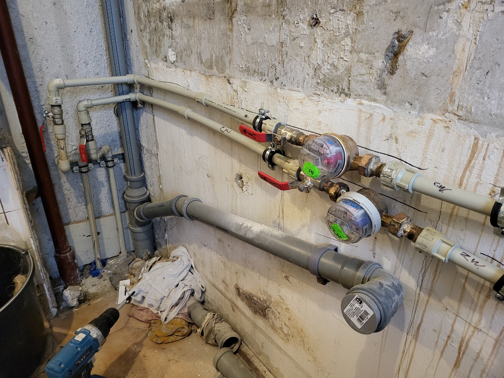 Montaż wodomierzy na rurach instalacji wodnej, widoczne liczniki z zielonymi naklejkami, zawory z czerwonymi rączkami, rury z tworzywa sztucznego, wiertarka akumulatorowa i elementy instalacji...