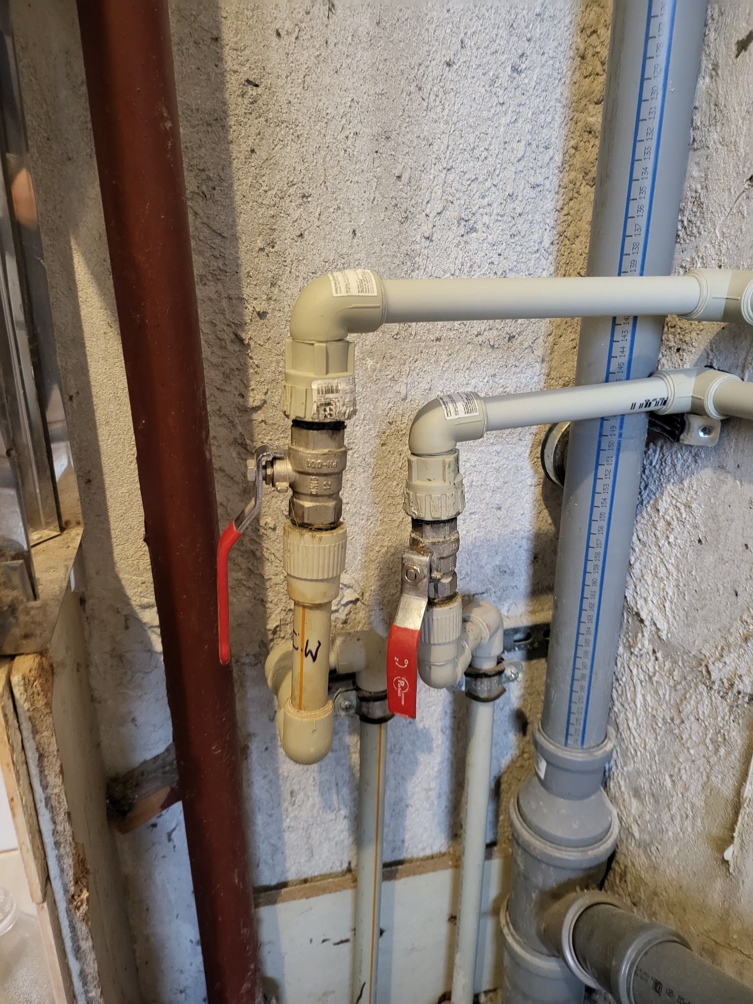 Fragment instalacji wodnej z rurami PCV, zaworami z czerwonymi rączkami i grubą rurą w kolorze rdzawym, zamontowanej przy szorstkiej ścianie.