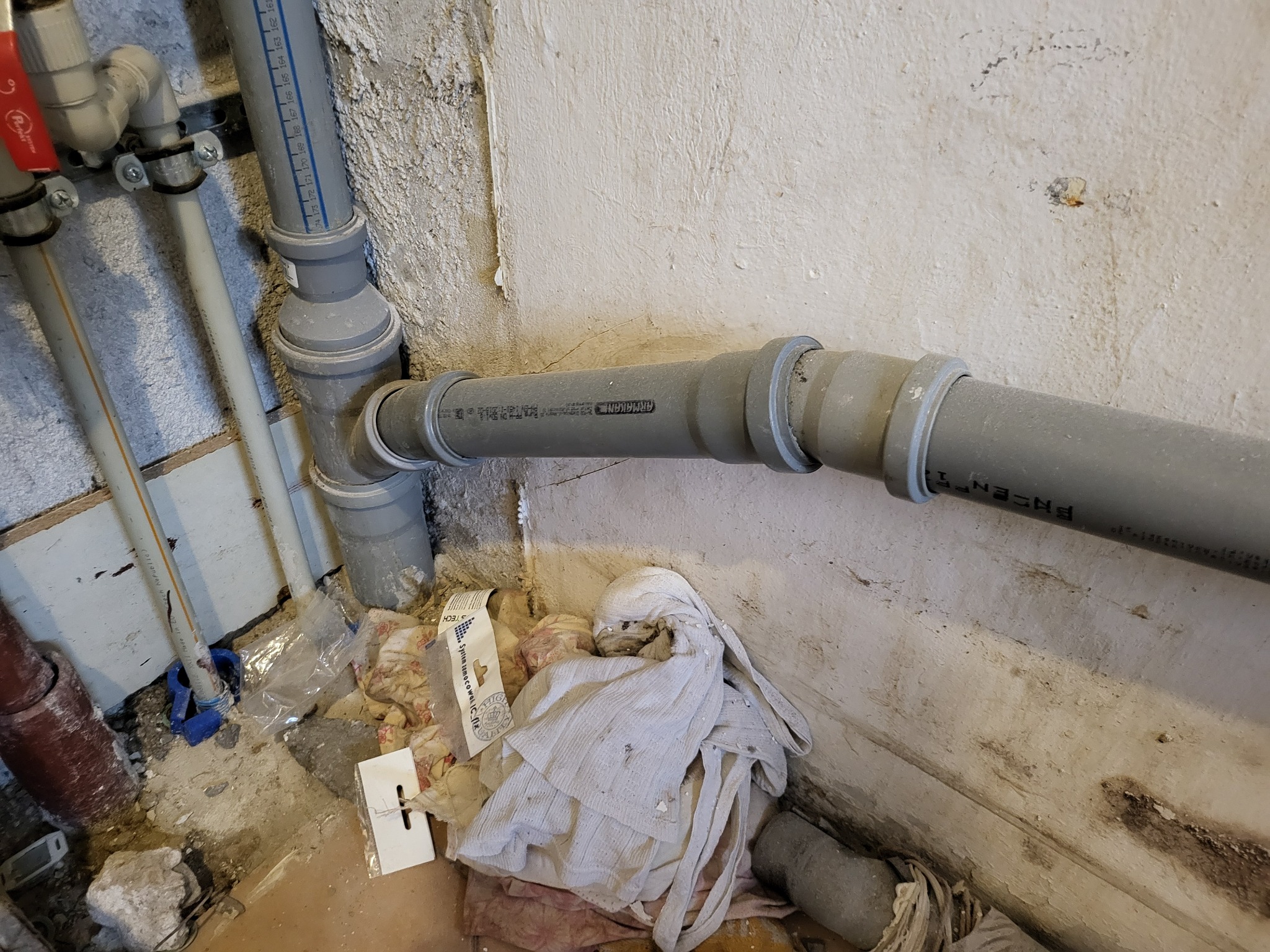 Szare rury kanalizacyjne z tworzywa sztucznego, połączone kolankiem, biegnące wzdłuż surowej, brudnej ściany, z widocznymi śladami po instalacji, na podłodze gruz, resztki materiałów budowlanych...