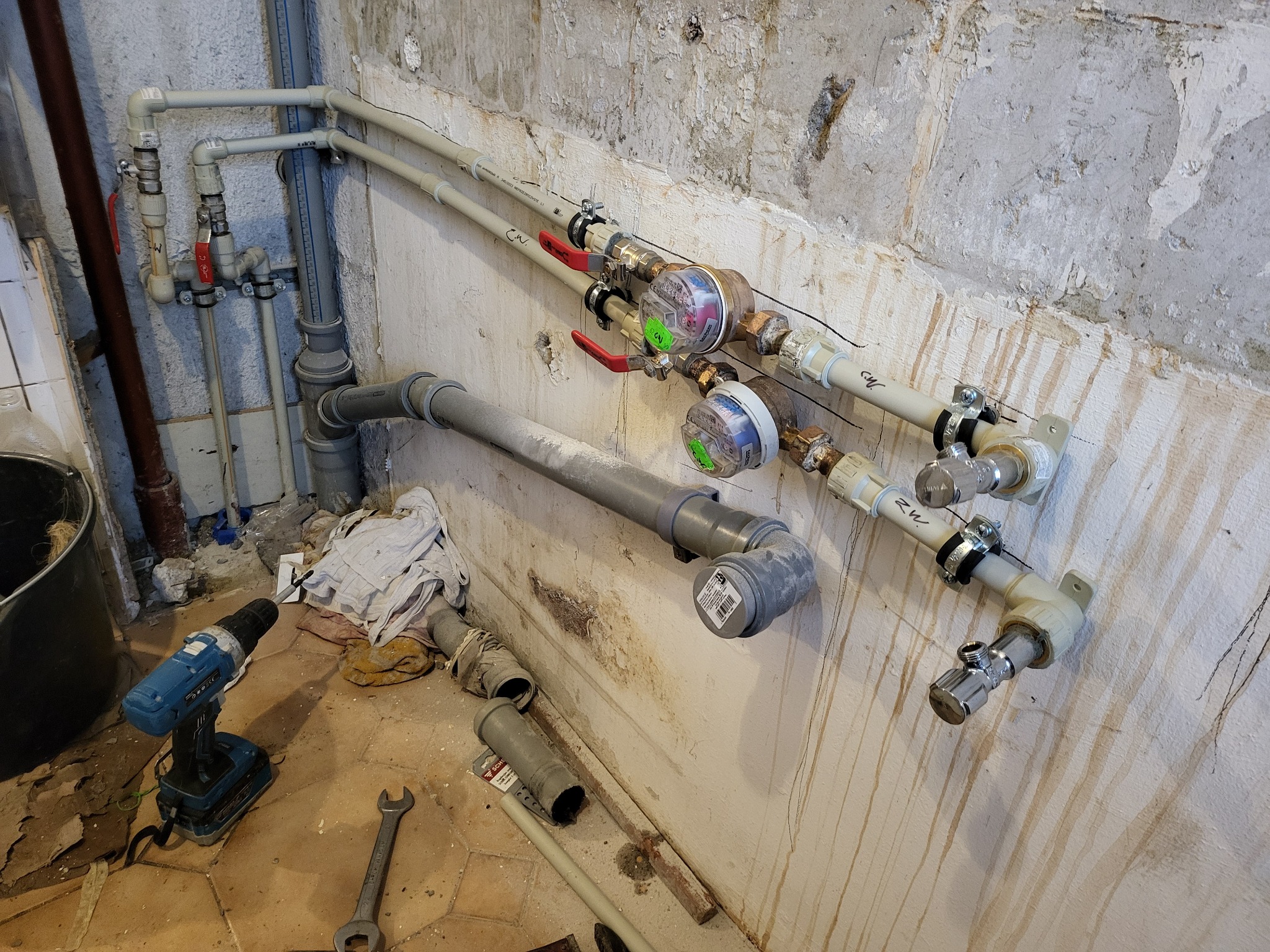 Zamontowane rury wodne z zaworami i licznikami na nierównej, odrapanej ścianie. Na podłodze wiertarka akumulatorowa i klucz płaski.