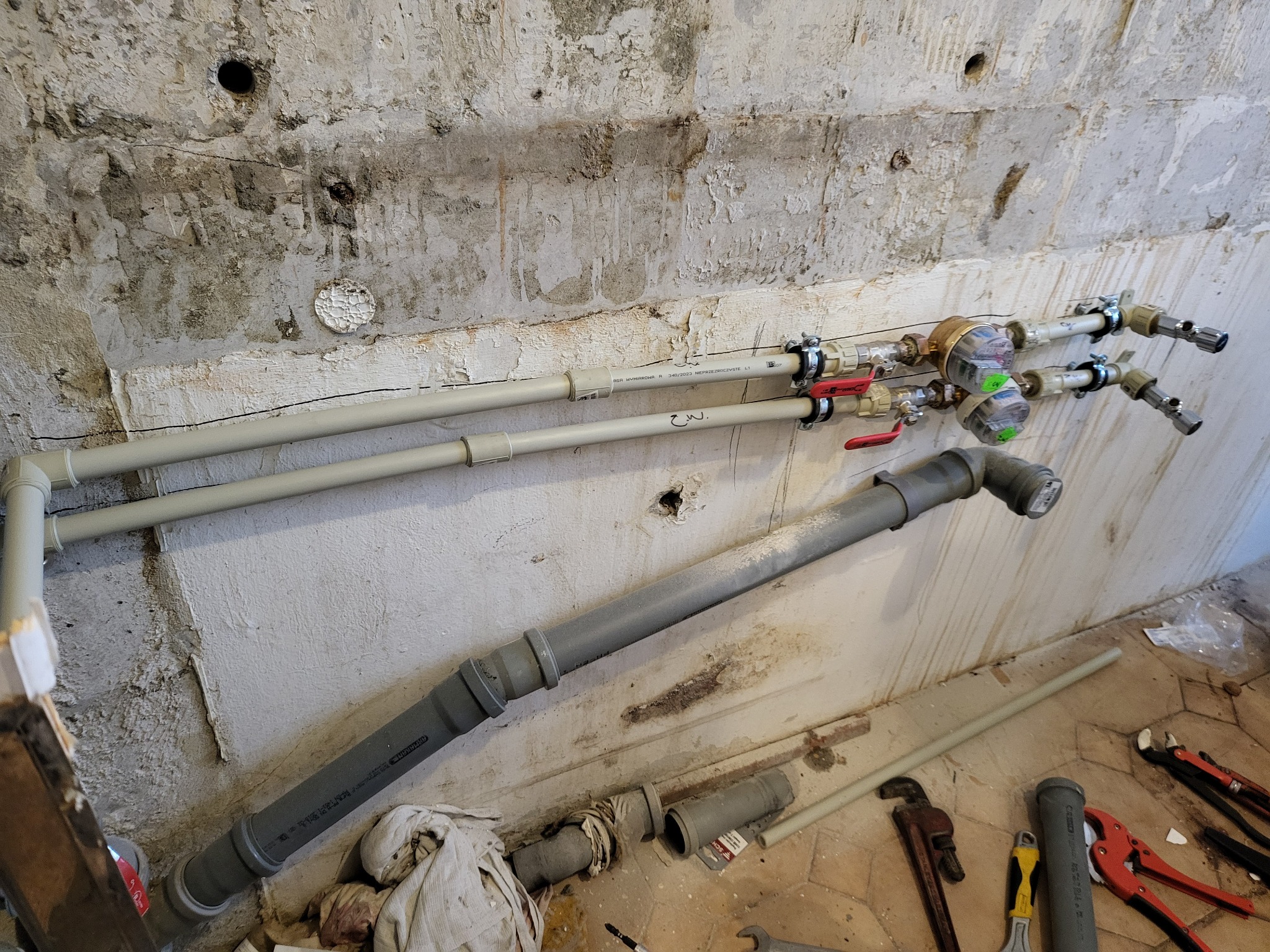 Zamontowane rury wodne z zaworami i licznikiem na tle surowej ściany z ubytkami tynku, widoczne narzędzia hydrauliczne na podłodze.