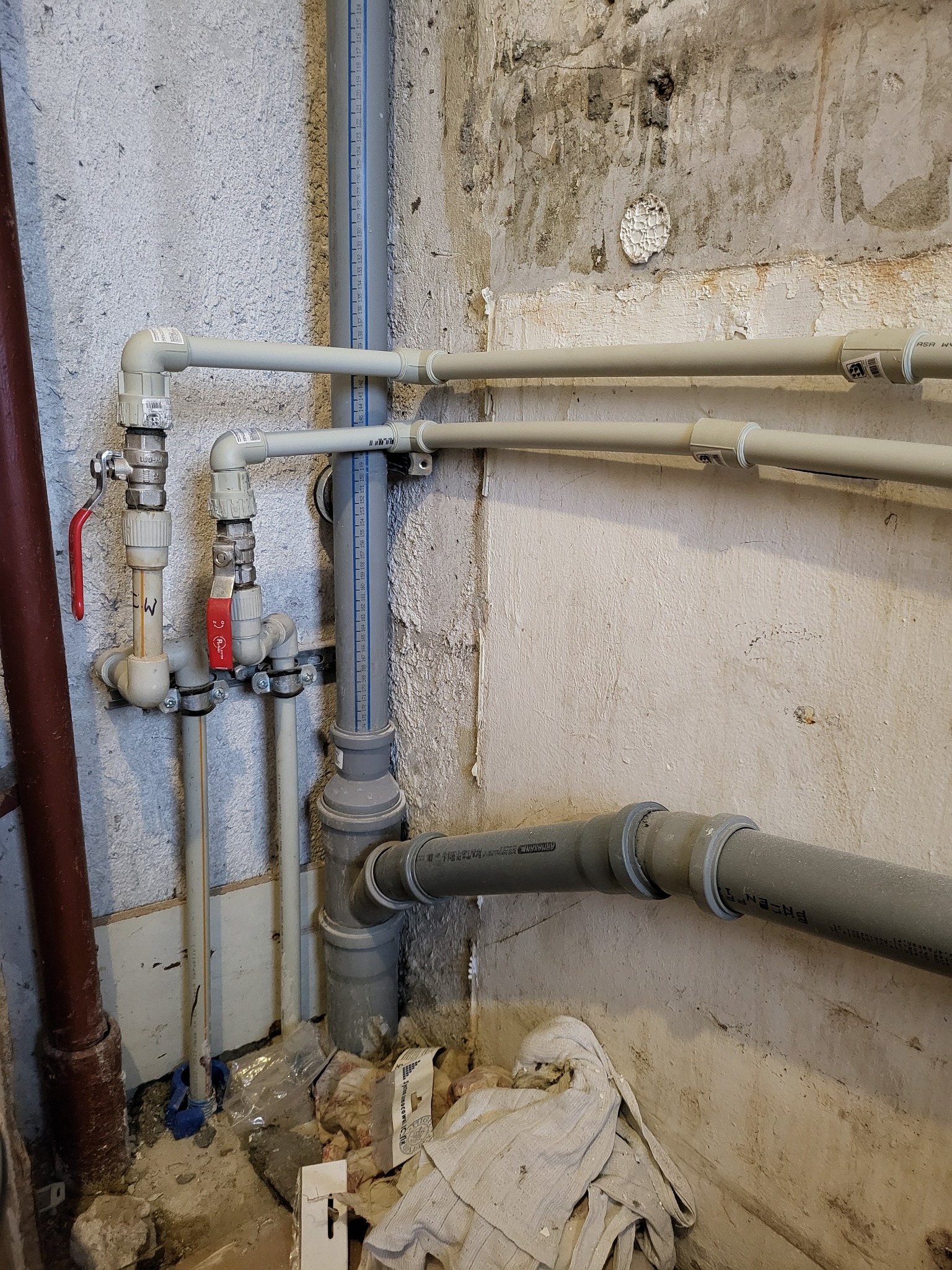 Szare rury instalacyjne z zaworami i oznaczeniami na tle surowej ściany z widocznymi śladami napraw, fragment instalacji wodnej.