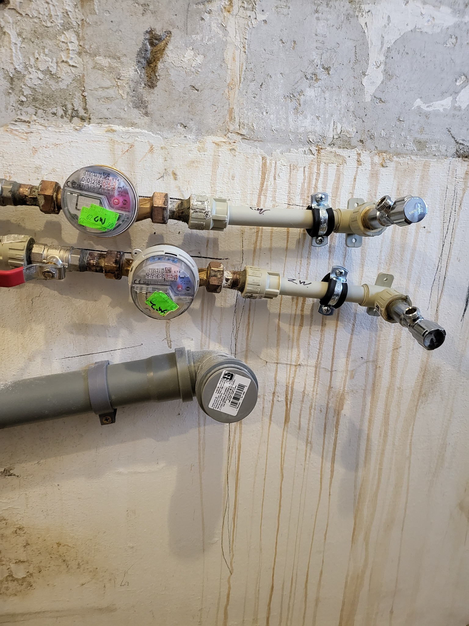Liczniki wody zamontowane na ścianie w Warszawie, z widocznymi rurami i zaworami, na tle ściany z odpryskującą farbą i zaciekami.