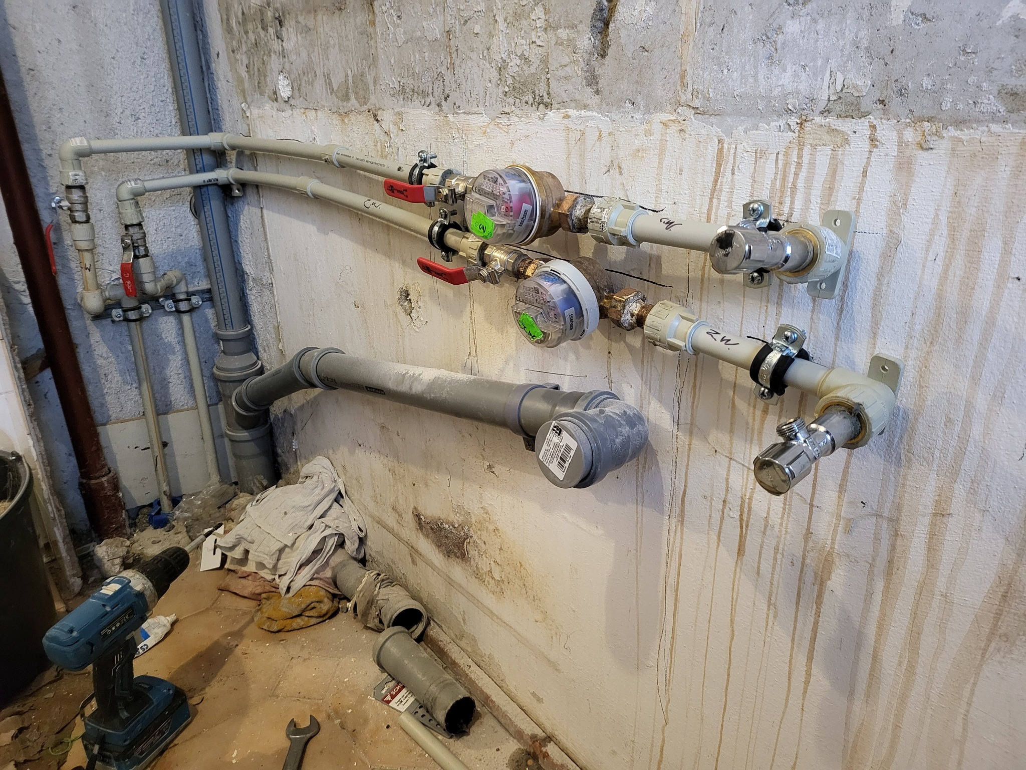 Dwa wodomierze zamontowane na rurach przy ścianie z zaciekami, obok wiertarka akumulatorowa i fragment rury kanalizacyjnej.