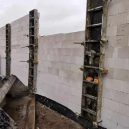 Ściana z białych bloczków silikatowych w trakcie murowania, wzmocniona szalunkiem systemowym. Na ścianie widoczne odręczne notatki. W tle palety z materiałami budowlanymi i częściowo widoczny budynek.