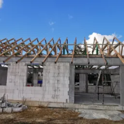Wznoszenie ścian z betonu komórkowego na budowie domu jednorodzinnego, widoczne drewniane elementy konstrukcji dachu i drabina oparta o ścianę.