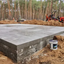 Świeżo wylana, gładka płyta fundamentowa z widoczną ceglaną podmurówką na piaszczystym gruncie, w tle las sosnowy i czerwony minikoparka.