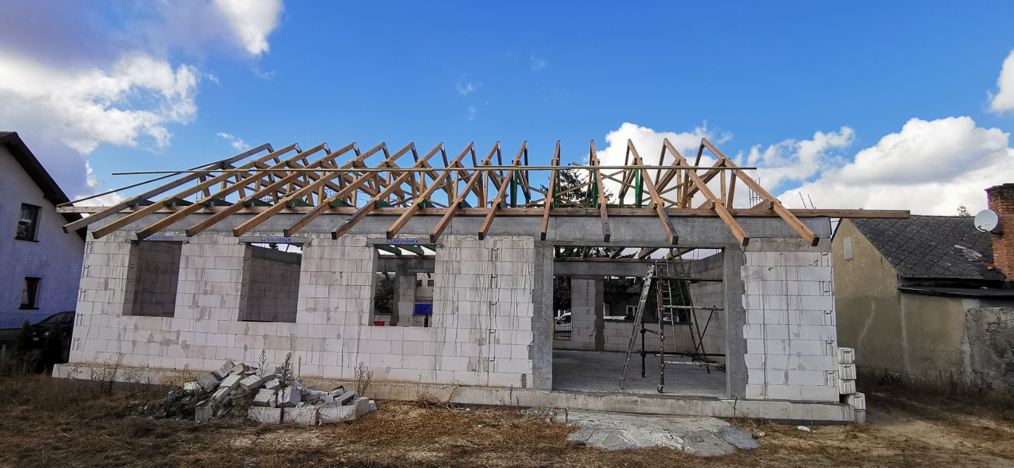 Wznoszenie ścian z betonu komórkowego na budowie domu jednorodzinnego, widoczne drewniane elementy konstrukcji dachu i drabina oparta o ścianę.