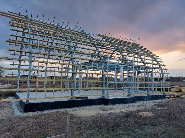 Konstrukcja szkieletowa budynku w trakcie montażu na betonowej płycie fundamentowej, widoczne elementy metalowe i drewniane, tło: zachmurzone niebo o zmierzchu.