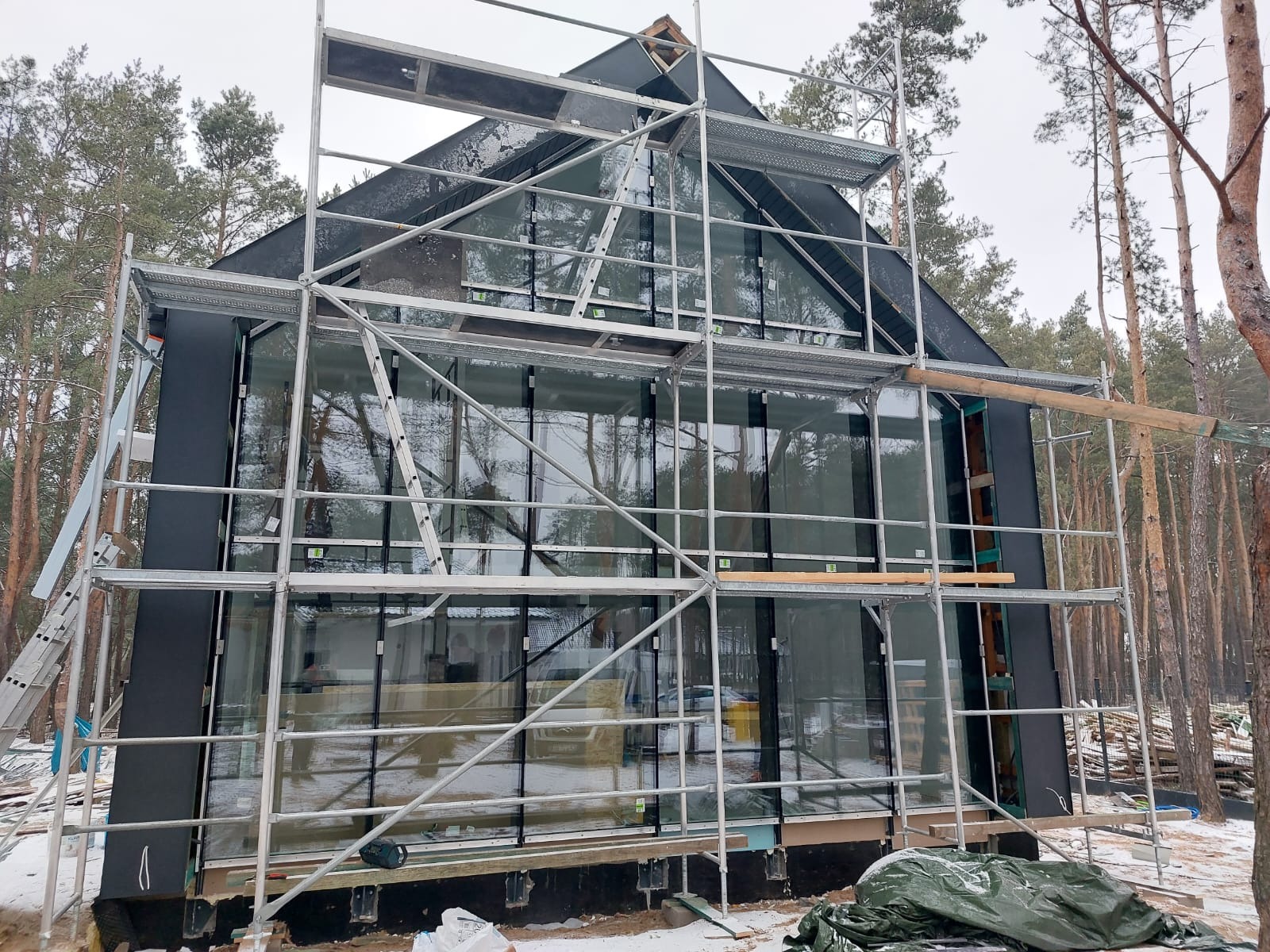 Dom szkieletowy w trakcie budowy, widoczne rusztowania i duże okna, tło stanowi las iglasty.