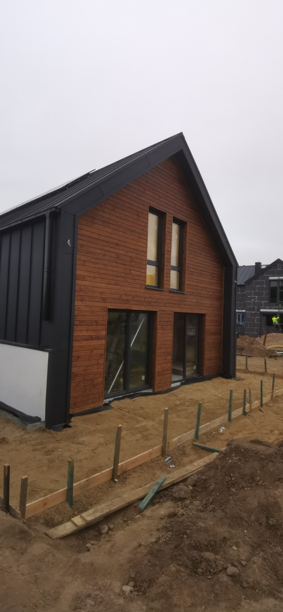 Nowoczesny dom z drewnianą elewacją i czarnymi elementami wykończeniowymi, widoczny w trakcie budowy, z przygotowanym terenem pod trawnik i widocznymi drewnianymi szalunkami.
