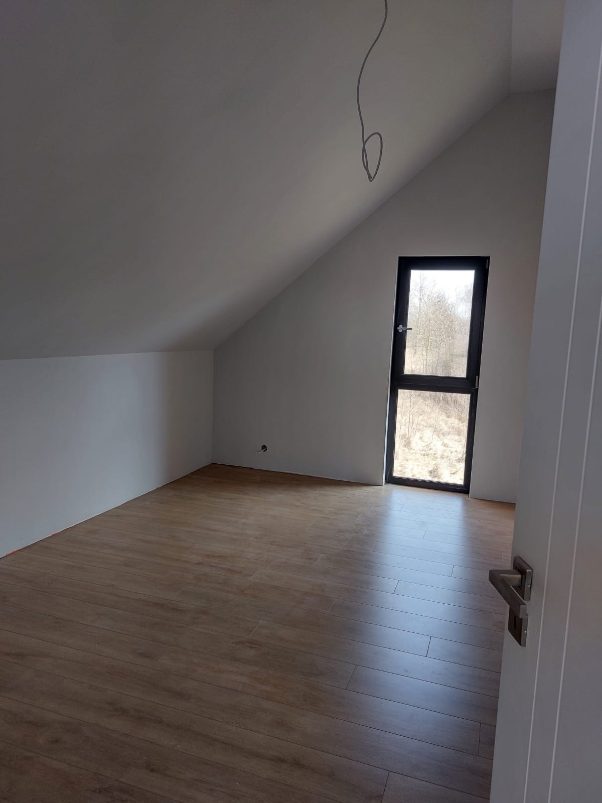 Poddasze w stanie deweloperskim z drewnopodobną podłogą, oknem balkonowym z czarną ramą i widocznym kablem zwisającym z sufitu.
