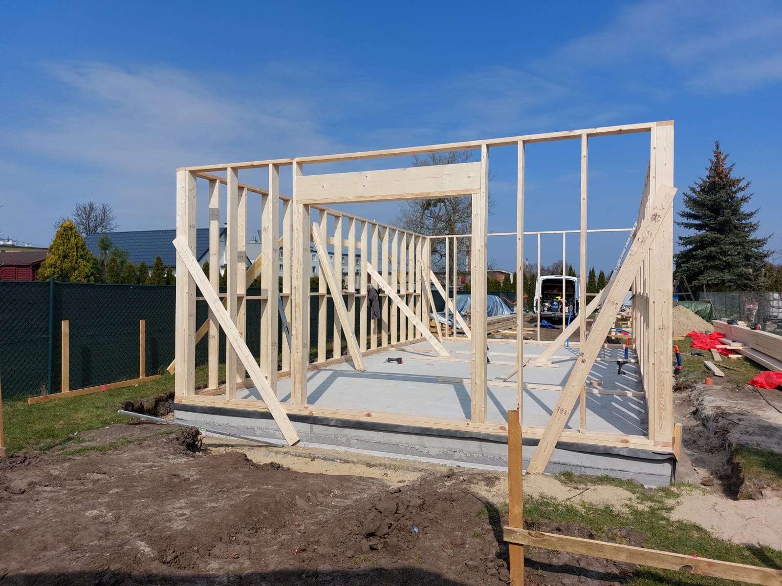 Wznoszenie drewnianej konstrukcji szkieletowej domu na betonowej płycie fundamentowej, widoczne deski i słupy tworzące szkielet, w tle zaparkowany biały van.