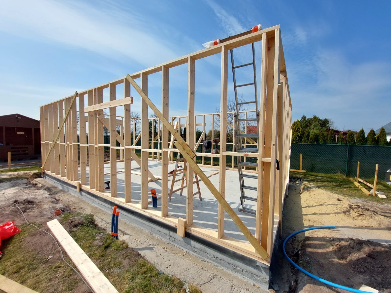 Wznoszenie drewnianej konstrukcji szkieletowej domu na betonowej płycie fundamentowej, widoczne drabiny, narzędzia i materiały budowlane, błękitne niebo w tle.