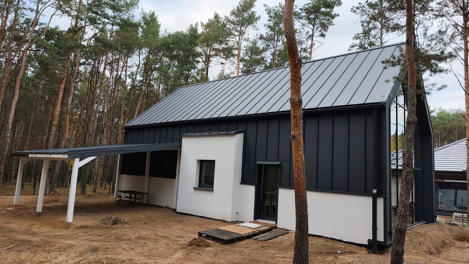 Nowoczesny dom szkieletowy z czarną elewacją i szarym dachem na tle lasu. Widoczny fragment tarasu i duże okno. Minimalistyczny design, naturalne otoczenie.