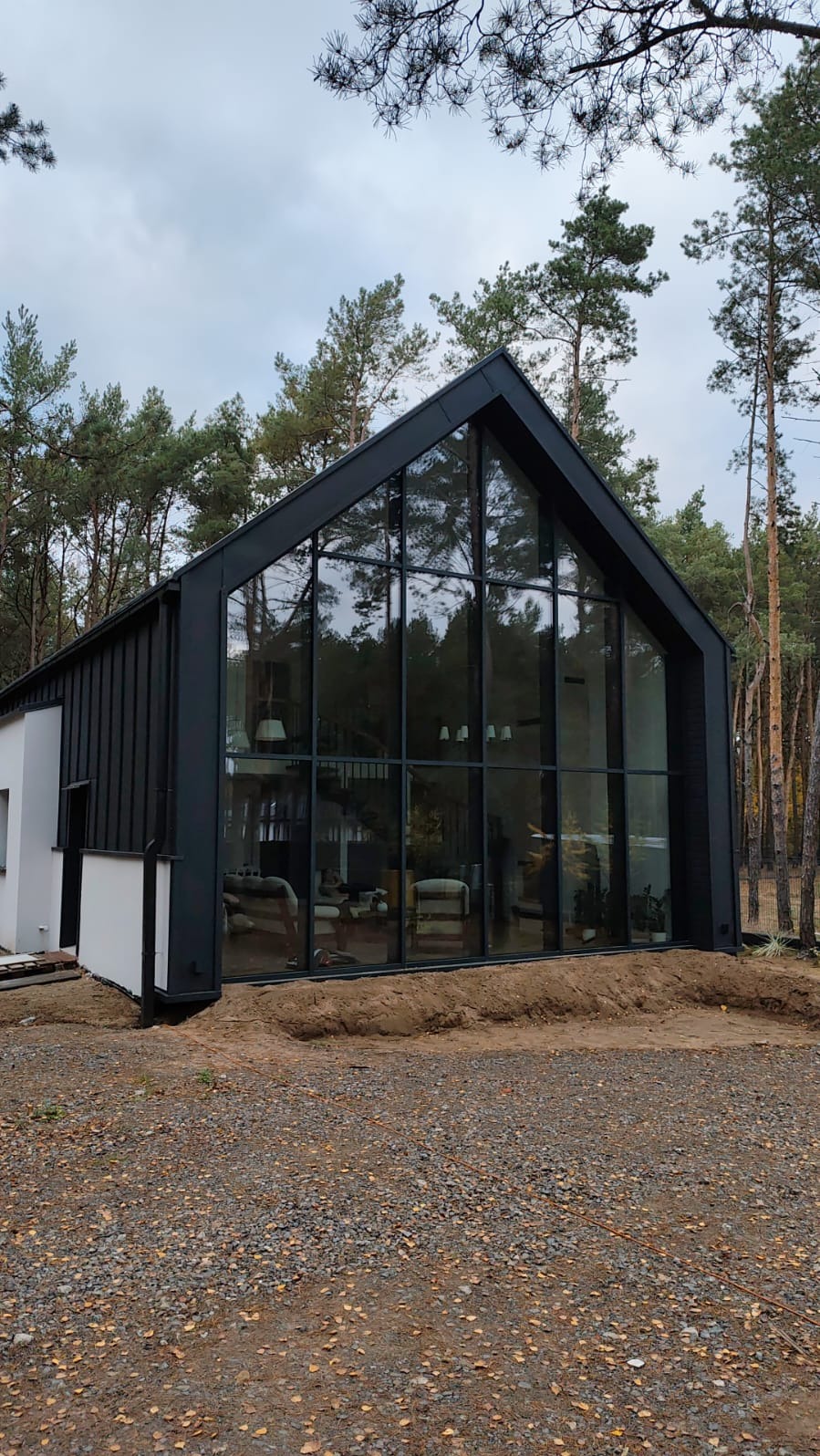 Nowoczesny dom szkieletowy z dużym przeszkleniem, widok z zewnątrz. Czarna elewacja kontrastuje z jasnym wnętrzem. Otoczenie leśne, ziemia pokryta żwirem.