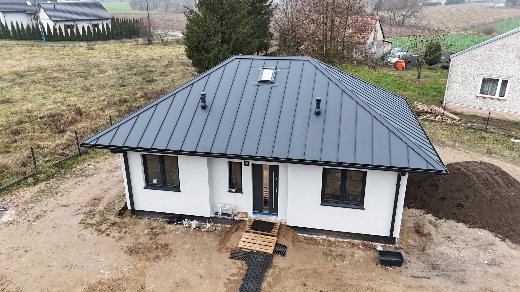 Nowoczesny, parterowy dom z ciemnoszarą, metalową blachą na dachu i białymi ścianami. Widok z góry, otoczenie w stanie surowym, palety przed wejściem.