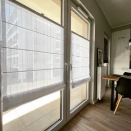 Jasne, półprzezroczyste osłony okienne w stylu rzymskim, zamontowane na dużym oknie balkonowym, z widokiem na budynek z balkonami i jasną podłogę balkonu.