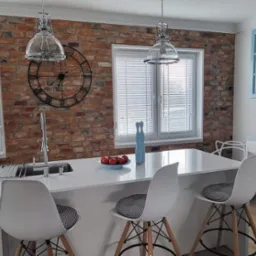 Jasna kuchnia z ceglaną ścianą, oknami z białymi żaluzjami poziomymi i wyspą z białym blatem oraz krzesłami barowymi.