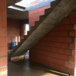 Wnętrze domu w trakcie budowy: murowane ściany z czerwonej cegły, surowy betonowy strop i tymczasowe, drewniane schody z widocznym zbrojeniem, w tle taczka budowlana.