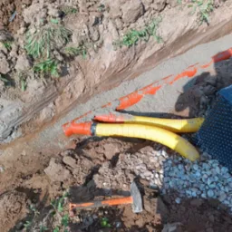 Wykop z podsypką piaskową, częściowo zasypane rury drenarskie w kolorze ż&oacute;łtym i pomarańczowym, widoczna folia kubełkowa i kamień drenażowy, obok leży młotek.