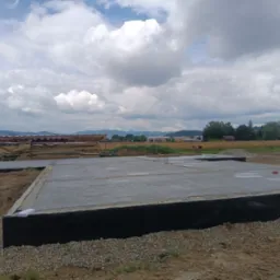 Zalany betonowy fundament z izolacją przeciwwilgociową, widok na otwarty teren z elementami drewnianymi w tle, pochmurne niebo.