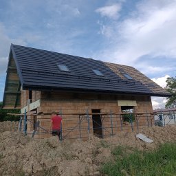 Marcin Kita - Dom w trakcie budowy z częściowo położonym, ciemnym dachem z oknami dachowymi; widoczne rusztowania i stos ziemi.