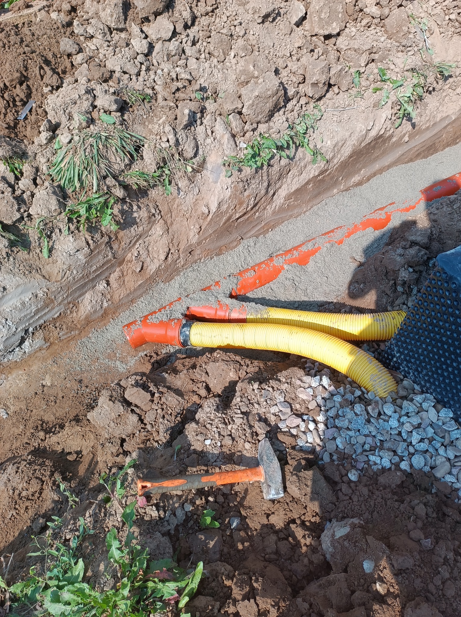 Wykop z podsypką piaskową, częściowo zasypane rury drenarskie w kolorze żółtym i pomarańczowym, widoczna folia kubełkowa i kamień drenażowy, obok leży młotek.
