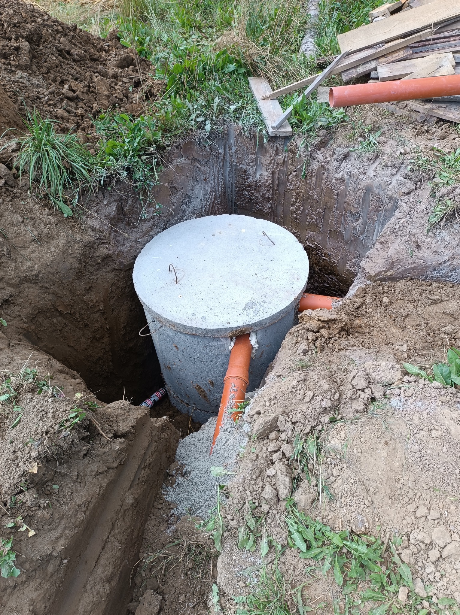 Betonowy zbiornik na szambo w wykopie z podłączonymi rurami kanalizacyjnymi, widoczne narzędzie uszczelniające i sterta ziemi z trawą.