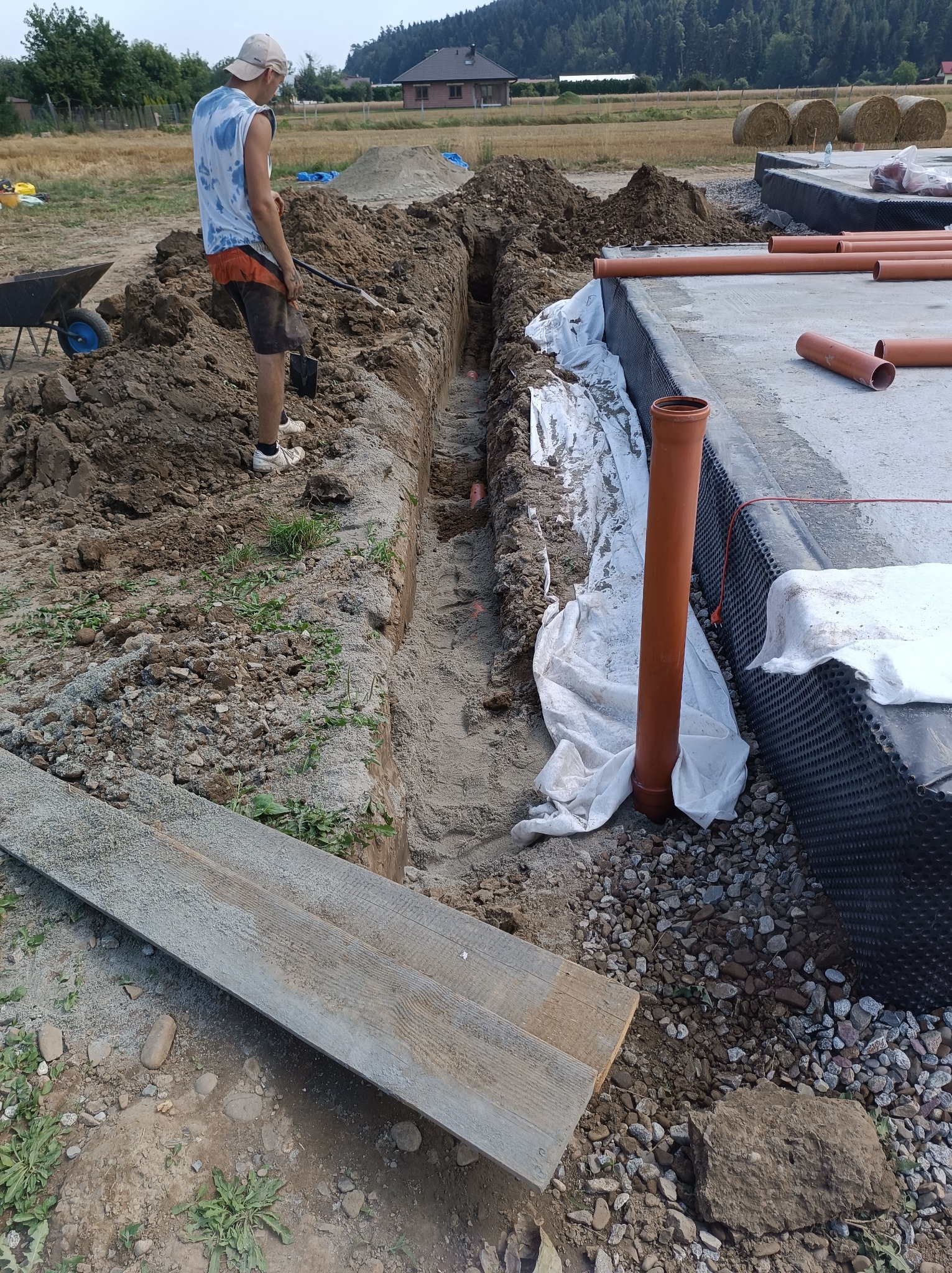 Prace ziemne przy budowie fundamentów: wykopy pod instalacje, widoczny fragment izolacji pionowej fundamentów, rury kanalizacyjne, pracownik z łopatą, w tle pole i las.