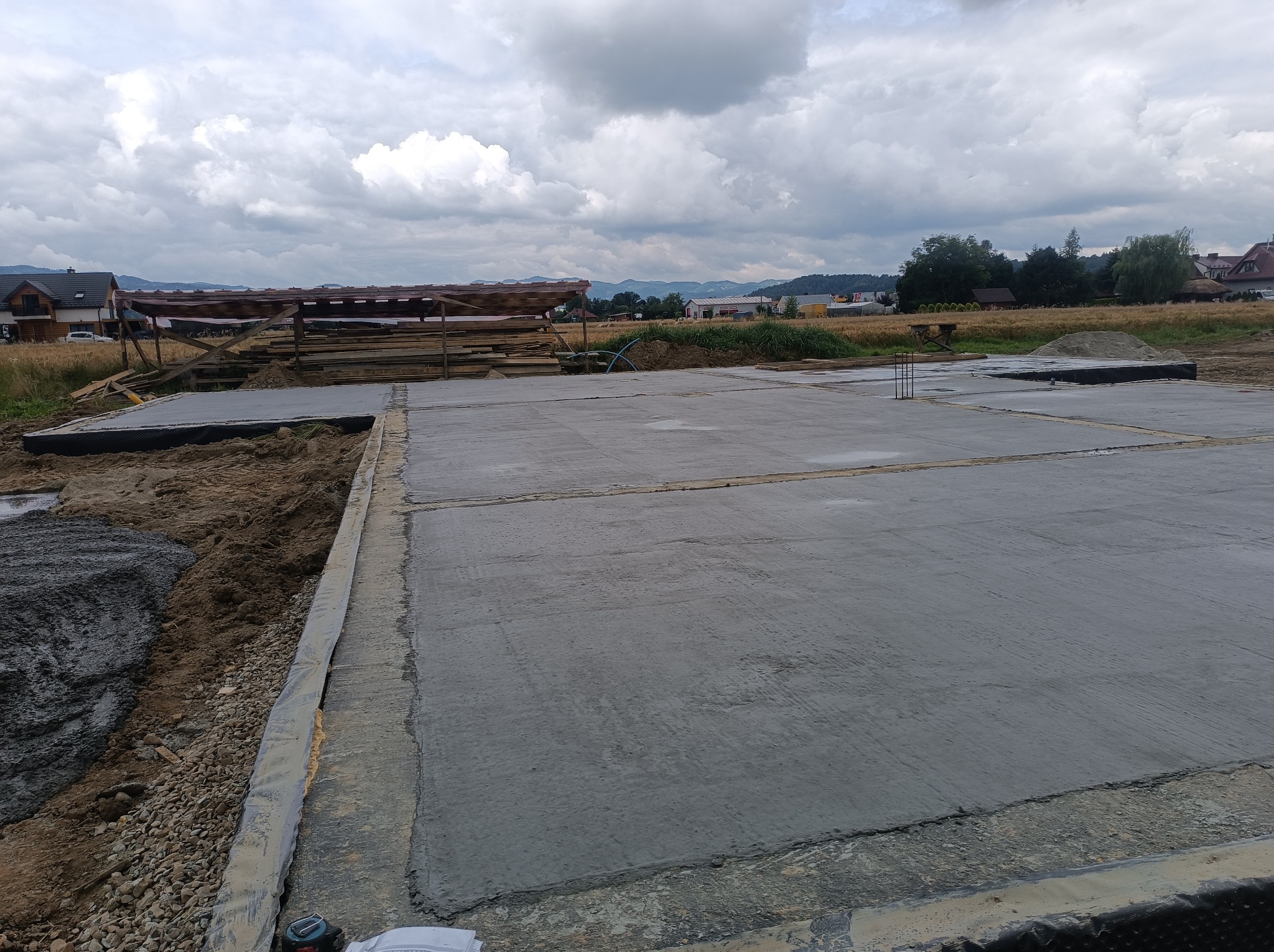 Zalana betonem płyta fundamentowa z izolacją pionową, widoczne wystające pręty zbrojeniowe, w tle teren budowy i pochmurne niebo.