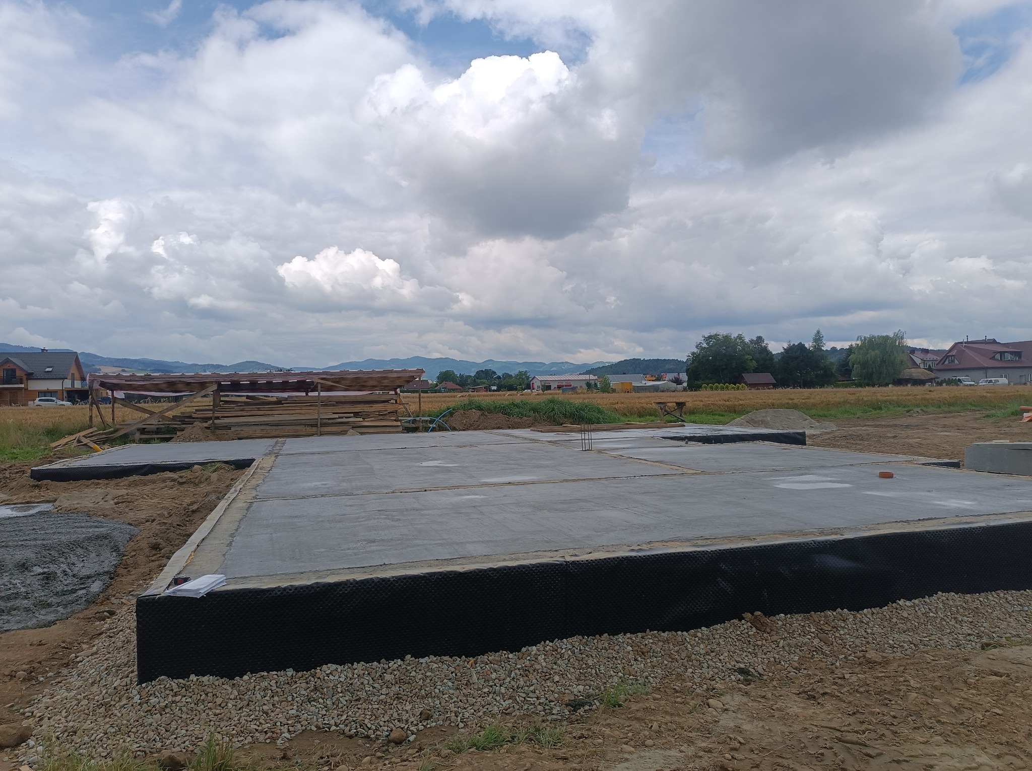 Zalany betonowy fundament z izolacją przeciwwilgociową, widok na otwarty teren z elementami drewnianymi w tle, pochmurne niebo.