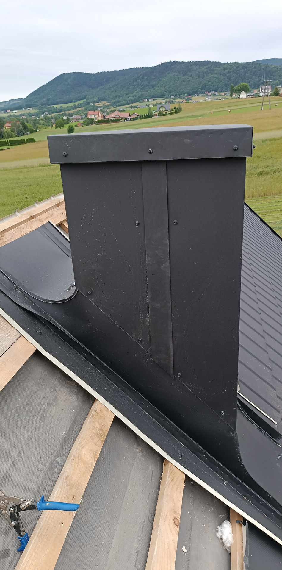 Czarny komin z blachy na dachu z blachy na tle zielonych wzgórz, widoczne drewniane elementy konstrukcji dachu i narzędzia dekarskie.