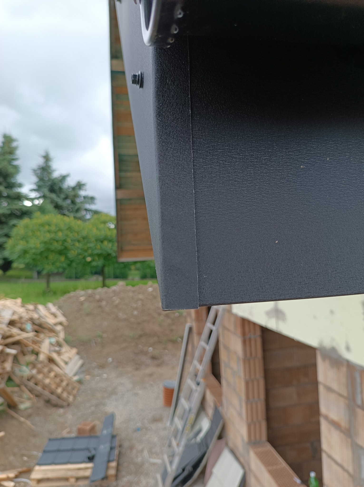 Fragment okapu dachu z blachy w kolorze grafitowym, widoczny na tle budowy domu z cegły ceramicznej i sterty drewna.