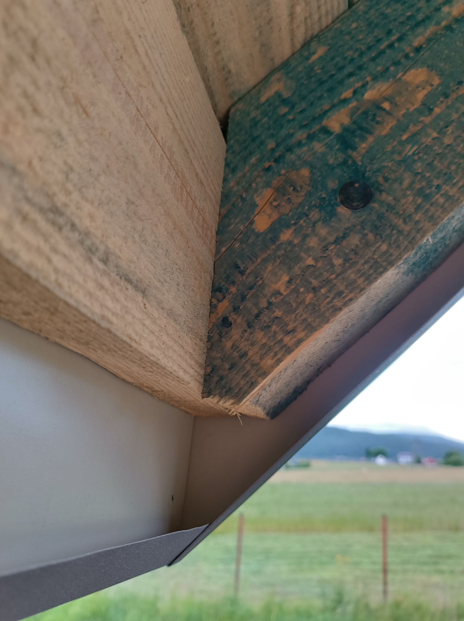 Fragment drewnianej konstrukcji dachu z widocznym elementem metalowej obróbki blacharskiej, na tle rozległego krajobrazu z polami i wzgórzami.