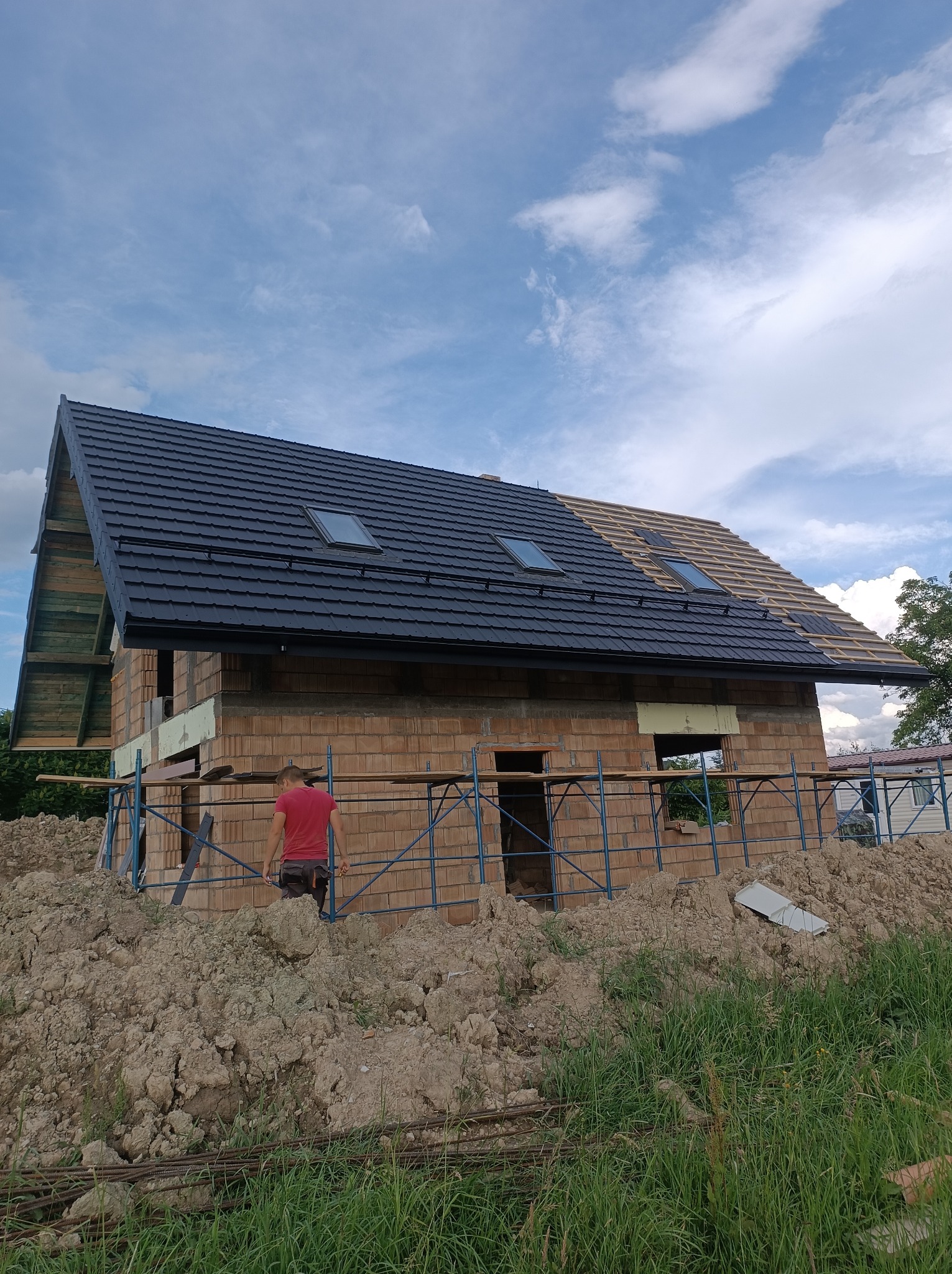 Dom w trakcie budowy z częściowo położonym, ciemnym dachem z oknami dachowymi; widoczne rusztowania i stos ziemi.